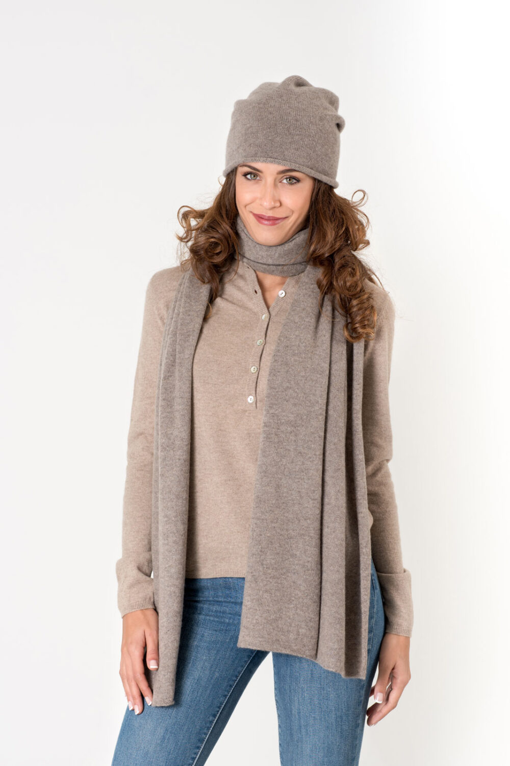 Donna sorridente con capelli ricci, indossa un maglione beige e una sciarpa dello stesso colore, in un ambiente neutro.