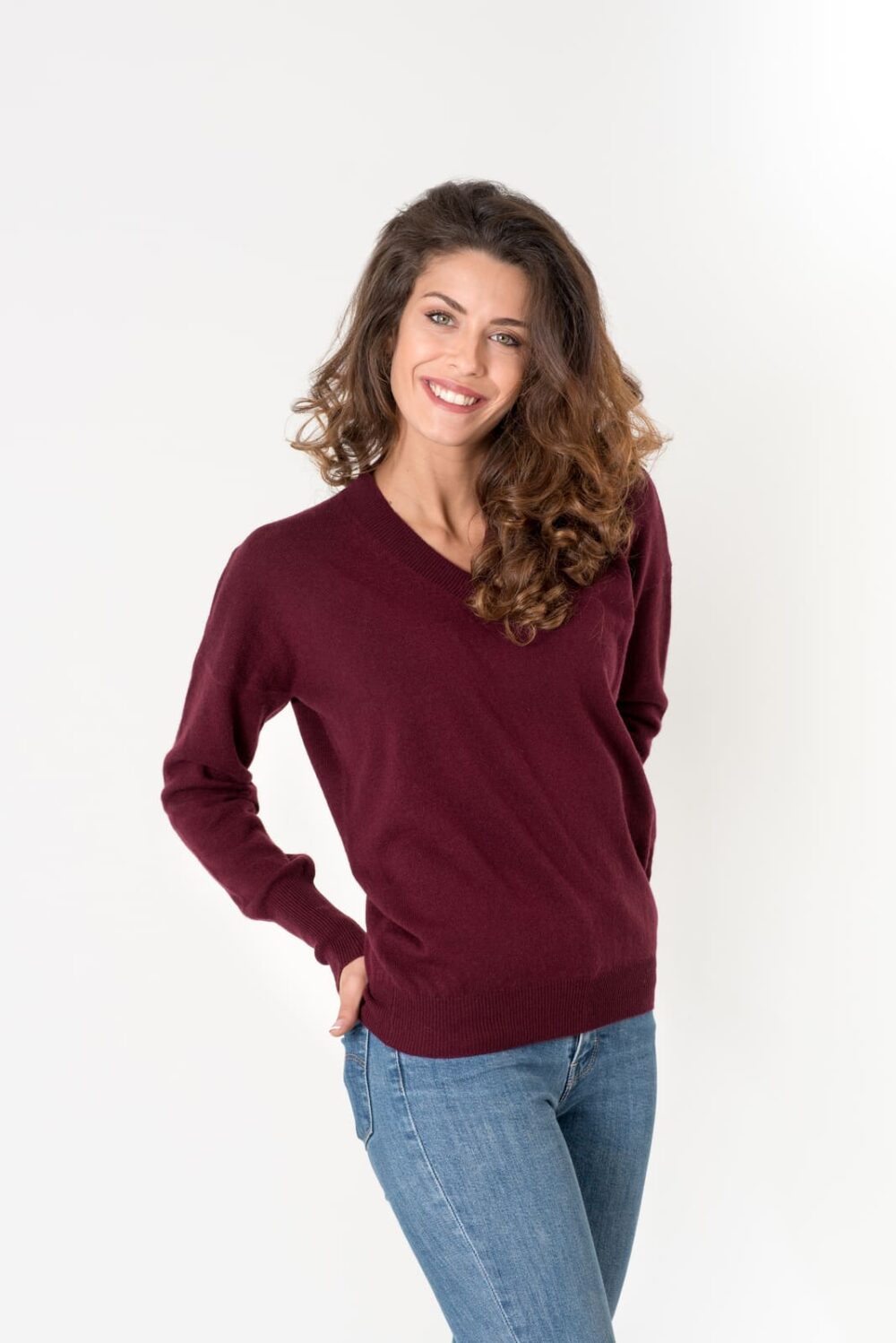 Ritratto di una giovane donna sorridente con capelli ricci, vestita con un maglione bordeaux a V, su sfondo bianco.